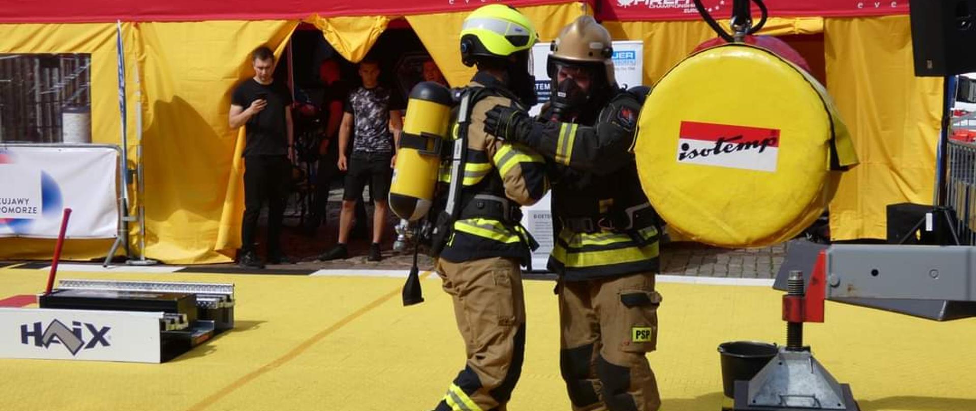 Na zdjęciu widać dwóch strażaków w umundurowaniu bojowym z butlami powietrznymi na plecach. Strażacy znajdują się na placu gdzie odbywają się zawody. W tle widać namioty. Strażacy stoją na żółtej macie. 