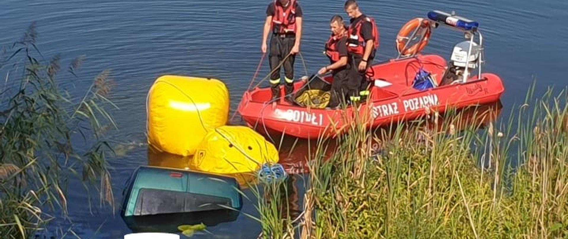 Zdjęcie przedstawia wynurzający się z otchłani stawu samochód osobowy do którego przymocowane są dwa balony wypornościowe oraz łódź motorową ze strażakami