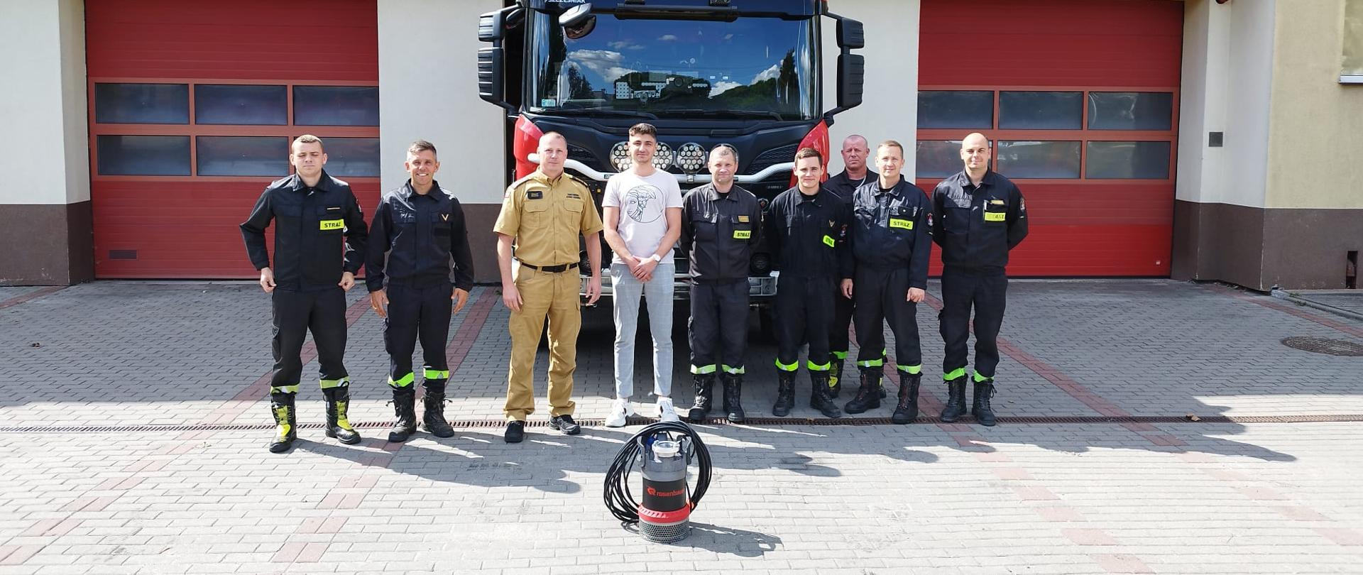 przed garażami wjazdowymi stoi auto pożarnicze. Przed autem stoi sześciu strażaków w mundurach koszarowych. W środku stoi komendant PSP i przedstawiciel firmy przekazujących pompę zanurzeniową.