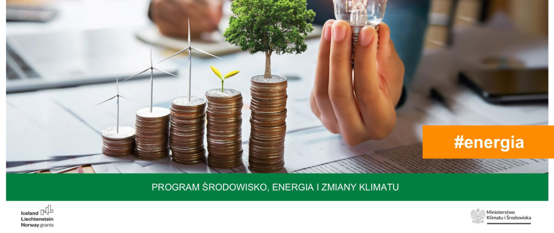 Stół, na nim dokumenty, na których leżą monety. Z nich wyrastają wiatraki, drzewo, roślina. Obok ręka mężczyzny trzymająca żarówkę. 