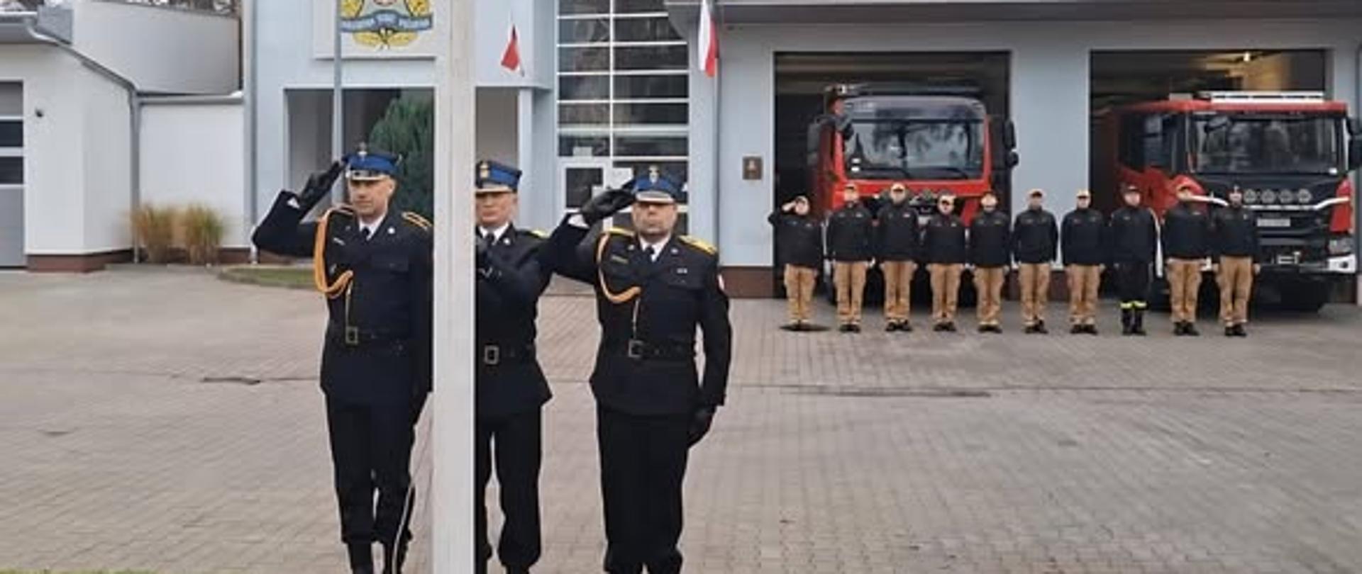 Poczet flagowy oddający honor. W tle strażacy na placu podczas uroczystej zbiórki.