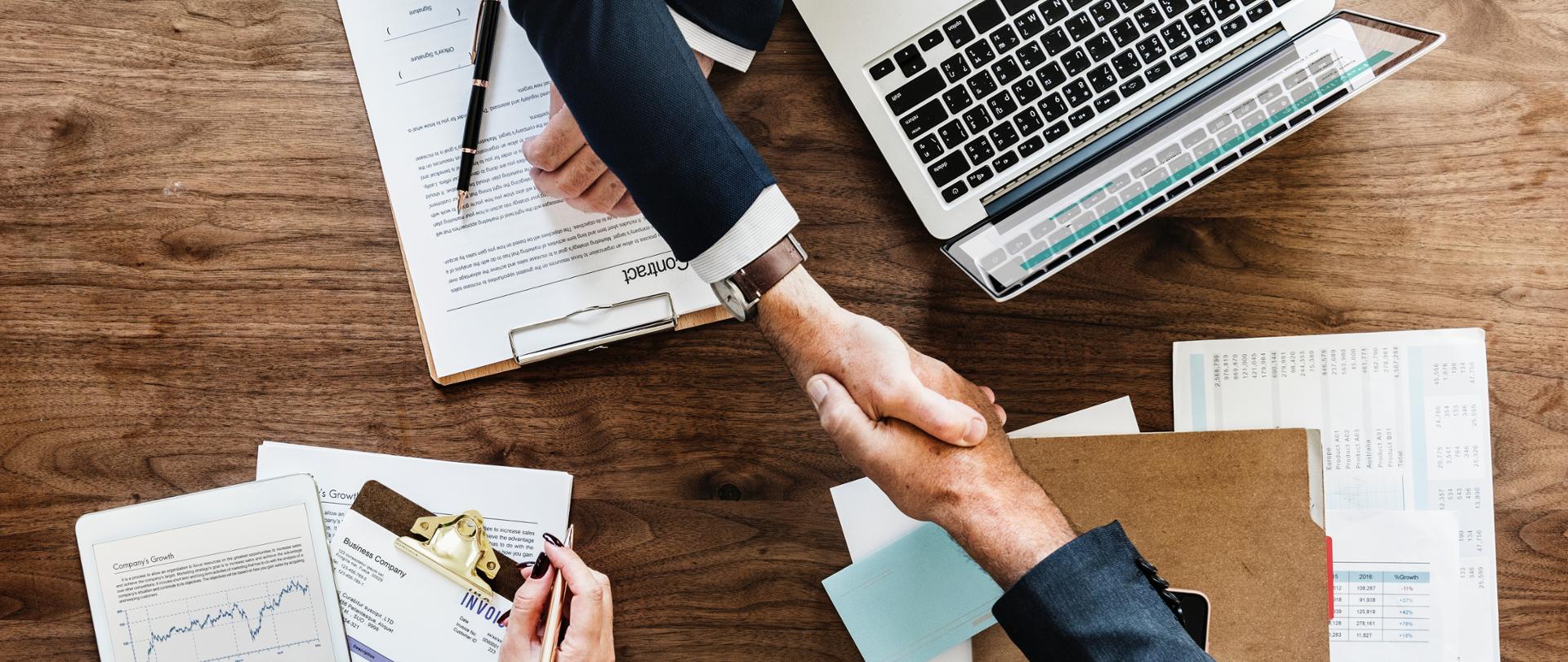 uścisk dłoni na tle macbooka oraz dokumentów