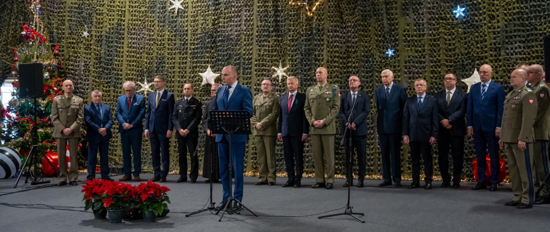 Na zdjęciu widać uroczystość w sali. Na środku mężczyzna w garniturze przemawia do mikrofonu, a za nim stoi rząd osób w mundurach wojskowych i garniturach. Tło stanowią świąteczne dekoracje: zielona siatka, świecące gwiazdy oraz choinka. Atmosfera jest oficjalna i uroczysta.