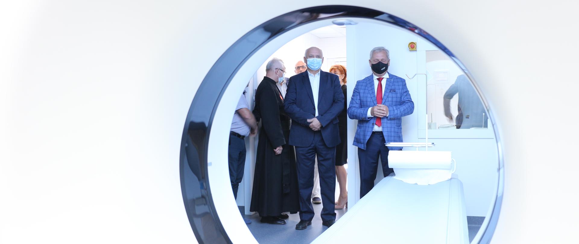 Zdjęcie prezentuje otwór aparatury do badania tomograficznego. Przez otwór, który znajduje się w centralnej części kadru, na dalszym planie w tle, widać uczestników uroczystego otwarcia pracowni tomograficznej. Wśród osób stojących w kadrze widać wojewodę opolskiego. Wojewoda trzyma swoje dłonie ułożone w pięść, na wysokości brzucha. Ubrany jest w jasny garnitur i czerwony krawat. Po jego prawej stronie stoi wysoki mężczyzna w średnim wieku. Spogląda w dal. Na przeciw wojewody widać leżankę - element wyposażenia pracowni tomografii.