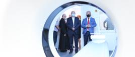 Zdjęcie prezentuje otwór aparatury do badania tomograficznego. Przez otwór, który znajduje się w centralnej części kadru, na dalszym planie w tle, widać uczestników uroczystego otwarcia pracowni tomograficznej. Wśród osób stojących w kadrze widać wojewodę opolskiego. Wojewoda trzyma swoje dłonie ułożone w pięść, na wysokości brzucha. Ubrany jest w jasny garnitur i czerwony krawat. Po jego prawej stronie stoi wysoki mężczyzna w średnim wieku. Spogląda w dal. Na przeciw wojewody widać leżankę - element wyposażenia pracowni tomografii.