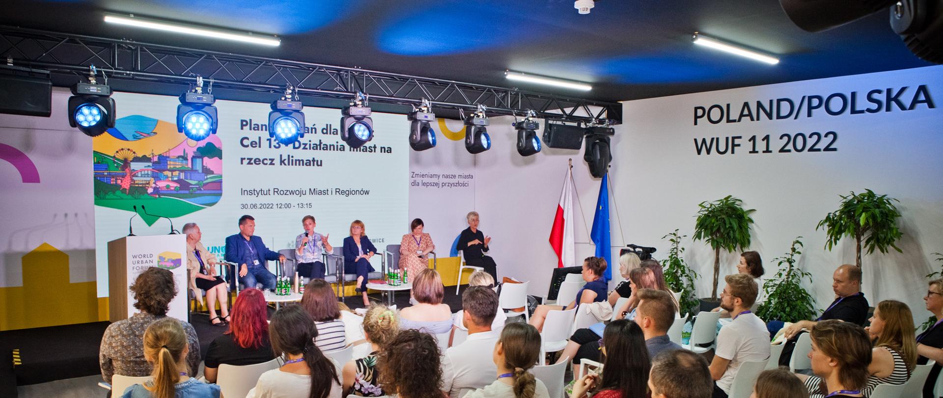Sesja podczas 11. Światowego Forum Miejskiego w 2022 roku. Na widowni osoby słuchają dyskusji między pięcioma osobami na podium.