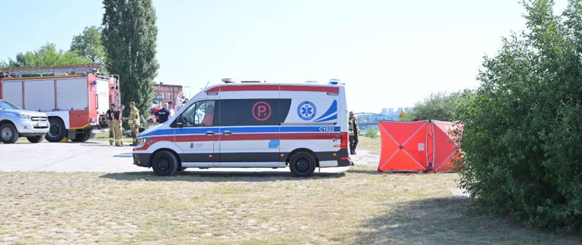 Zdjęcie przedstawia biało-czerwono-niebieską karetkę PRM. Obok niej stoi czerwony parawan osłaniający. W tle pojazdy straży pożarnej i Policji.