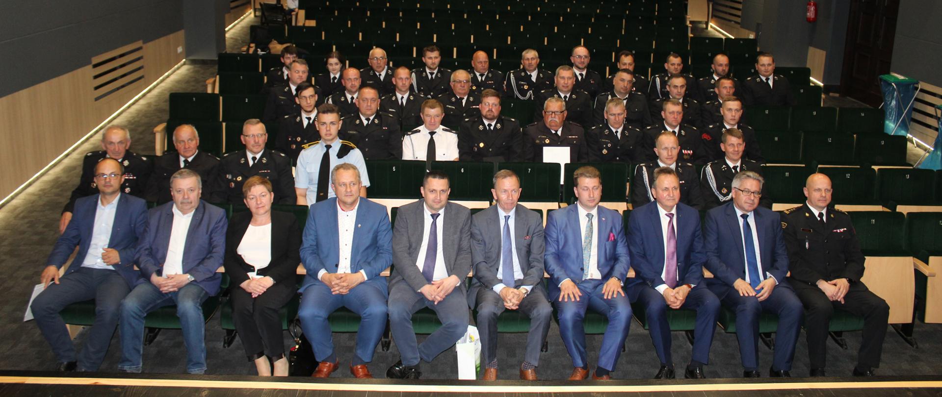 Strażacy ubrani w mundury galowe siedzą na krzesłach w czterech rzędach w sali kinowej. W pierwszym rzędzie siedzą posłowie na Sejm RP, komendant powiatowy PSP oraz przedstawiciele władz samorządowych. 