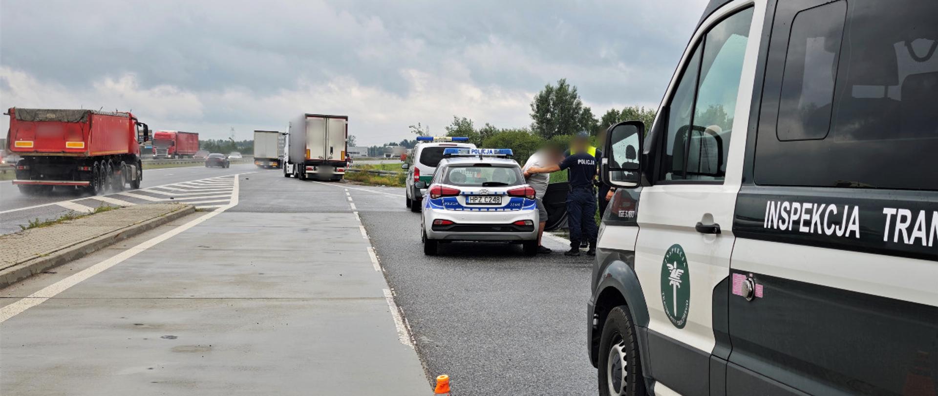 Na pierwszym planie (od prawej): przedni bok oznakowanego furgonu mazowieckiej Inspekcji Transportu Drogowego. W tle: za inspekcyjnym pojazdem służbowym stoi oznakowany radiowóz Policji. Policjanci umieszczają w radiowozie zatrzymanego kierowcę. Z policjantami rozmawia inspektor transportu drogowego. Na dalszym planie stoi zatrzymany do kontroli ciągnik siodłowy z naczepą.