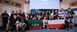 W dużej sali duża grupa ludzi, trzymają polską flagę i zielono-czarno-czerwoną flagę z napisem AGH, nad nimi na dużym ekranie napis ERC.