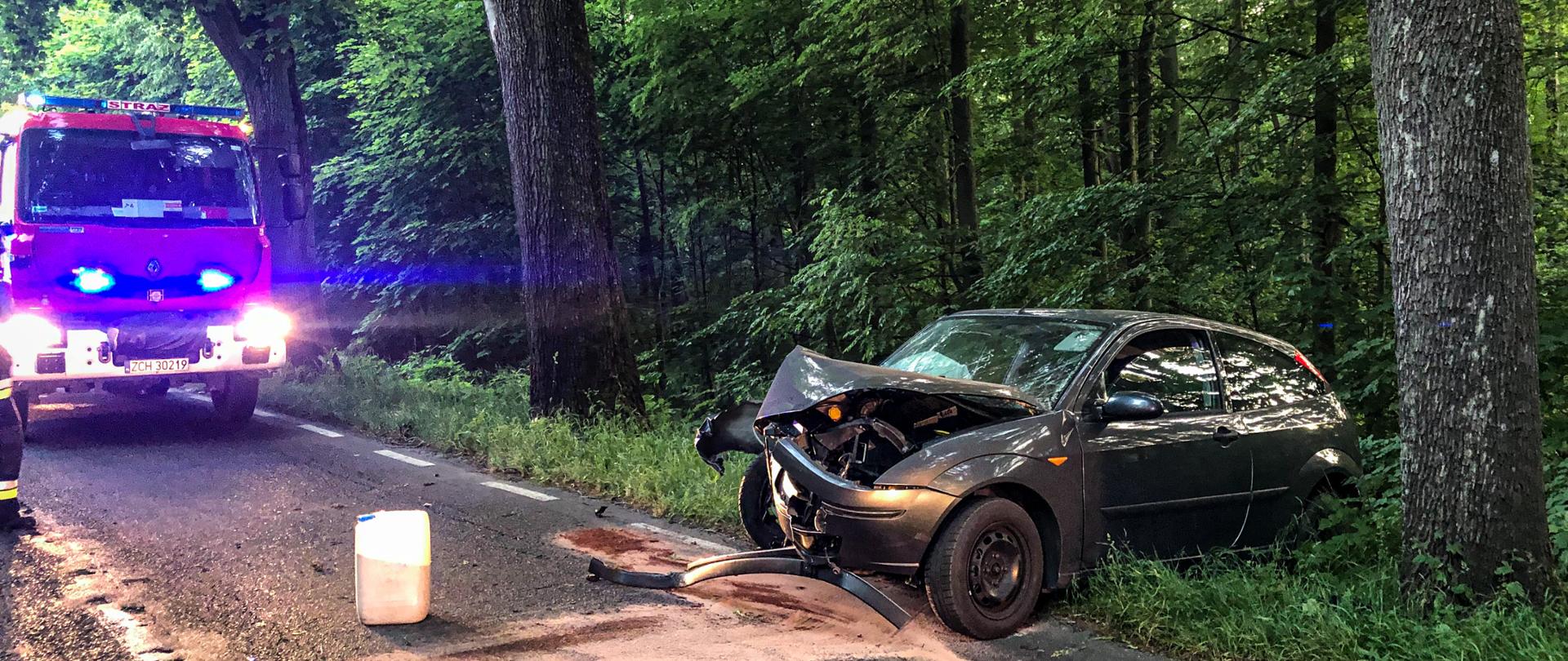 Na fotografii znajduje się rozbity samochód osobowy, który ma uszkodzony przód. Po lewej stronie na drodze stoi wóz strażacki, który ma uruchomione niebieskie sygnały świetlne.