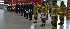 umundurowani strażacy stojący w garażu w dwuszeregu na tle auta strażackiego