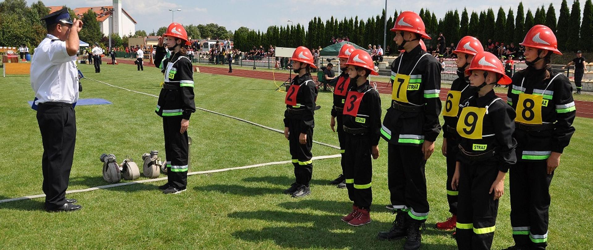 Na zdjęciu po lewej stronie sędzia zawodów przyjmujący meldunek od dowódcy młodzieżowej drużyny pożarniczej podczas wykonywania musztry