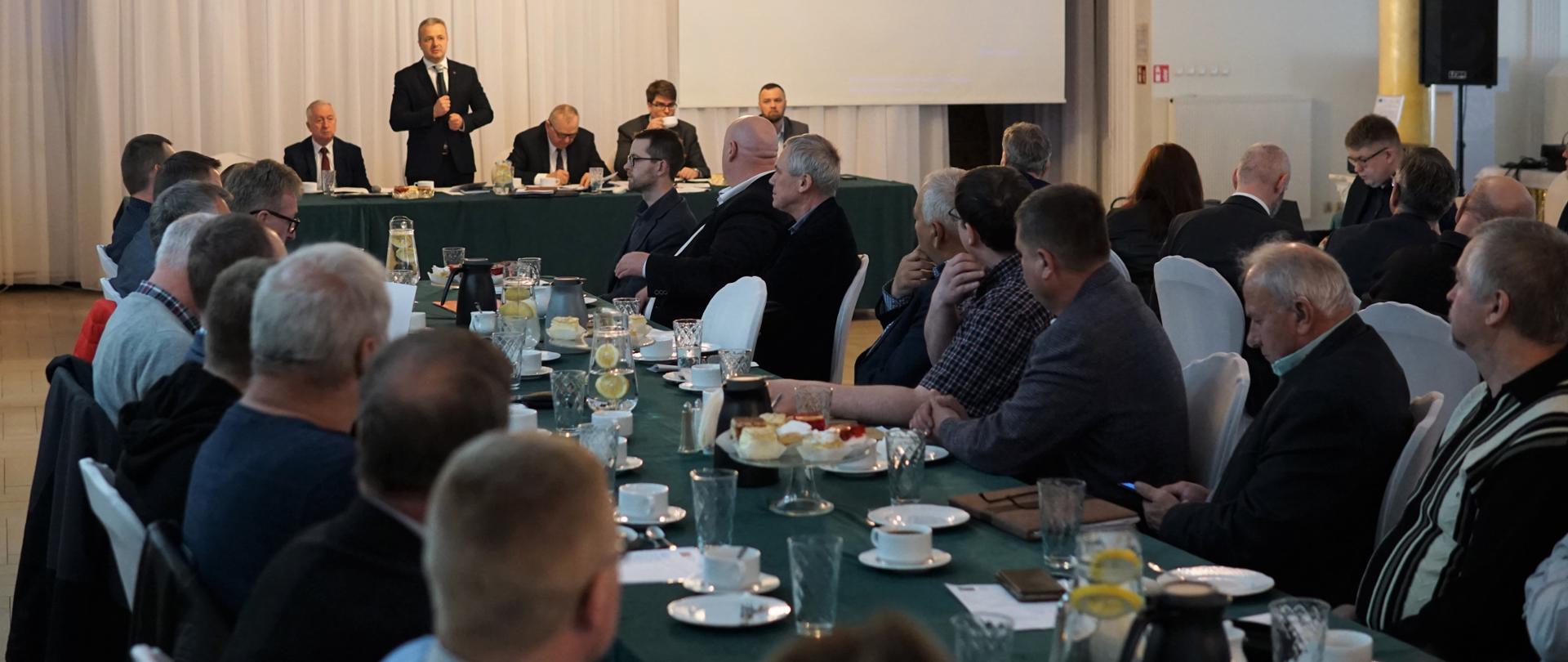 Jeden mężczyzna stojący z mikrofonem i wypowiadający się. Obok siedzi kilku mężczyzn