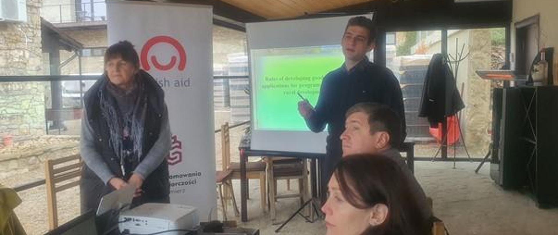 Grupa czterech osób uczestniczy w szkoleniu, w tle rollup z logo Polskiej pomocy oraz ekran i rzutnik multimedialny.