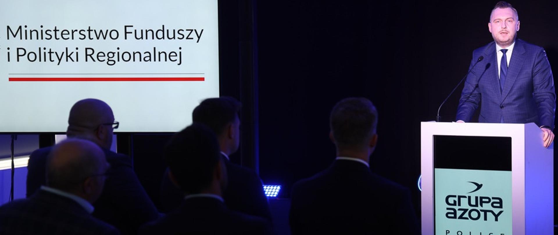 Wiceminister Konrad Wojnarowski w mównicy. Obok na ekranie napis: "Ministerstwo Funduszy i Polityki Regionalnej". Przed nim siedzi publiczność.