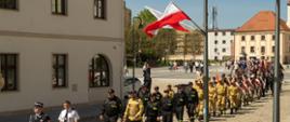 Fotografia wykonana z podwyższenia przedstawia uroczysty przemarsz kolumny strażaków oraz innych formacji, idących brukowaną ulicą w słoneczny dzień.