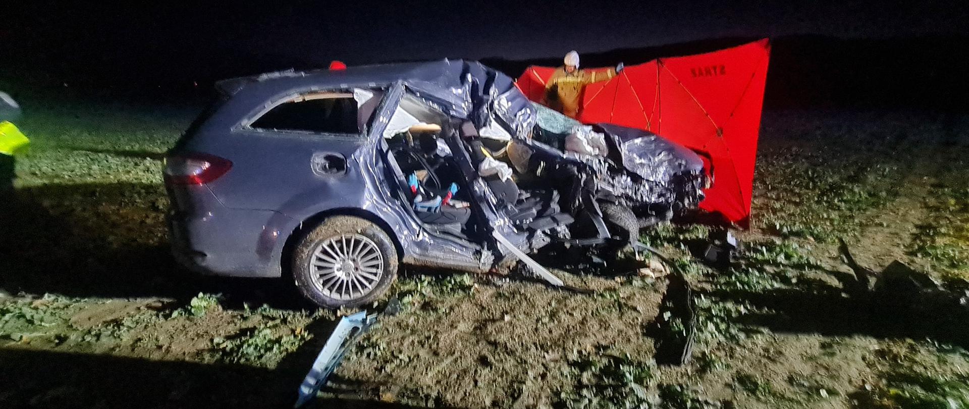 Obraz przedstawia rozbity pojazd osobowy. W tle strażak i rozłożony parawan. Pojazd w polu. Pora wieczorna.