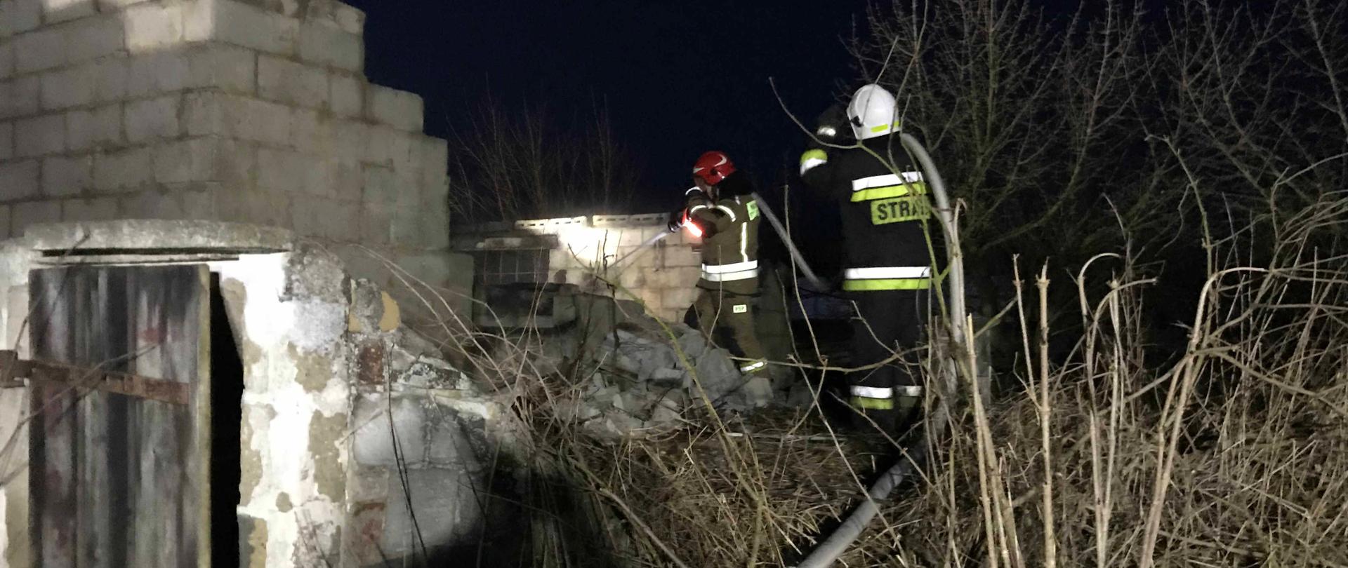 Zdjęcie przedstawia budynek gospodarczy. W środku zdjęcia widać dwóch strażaków podających jeden prąd wody w natarciu.
