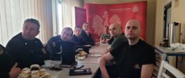 Osoby siedzą przy stole konferencyjnym. w Tle ścianka firmowa Państwowej Straż Pożarnej. 