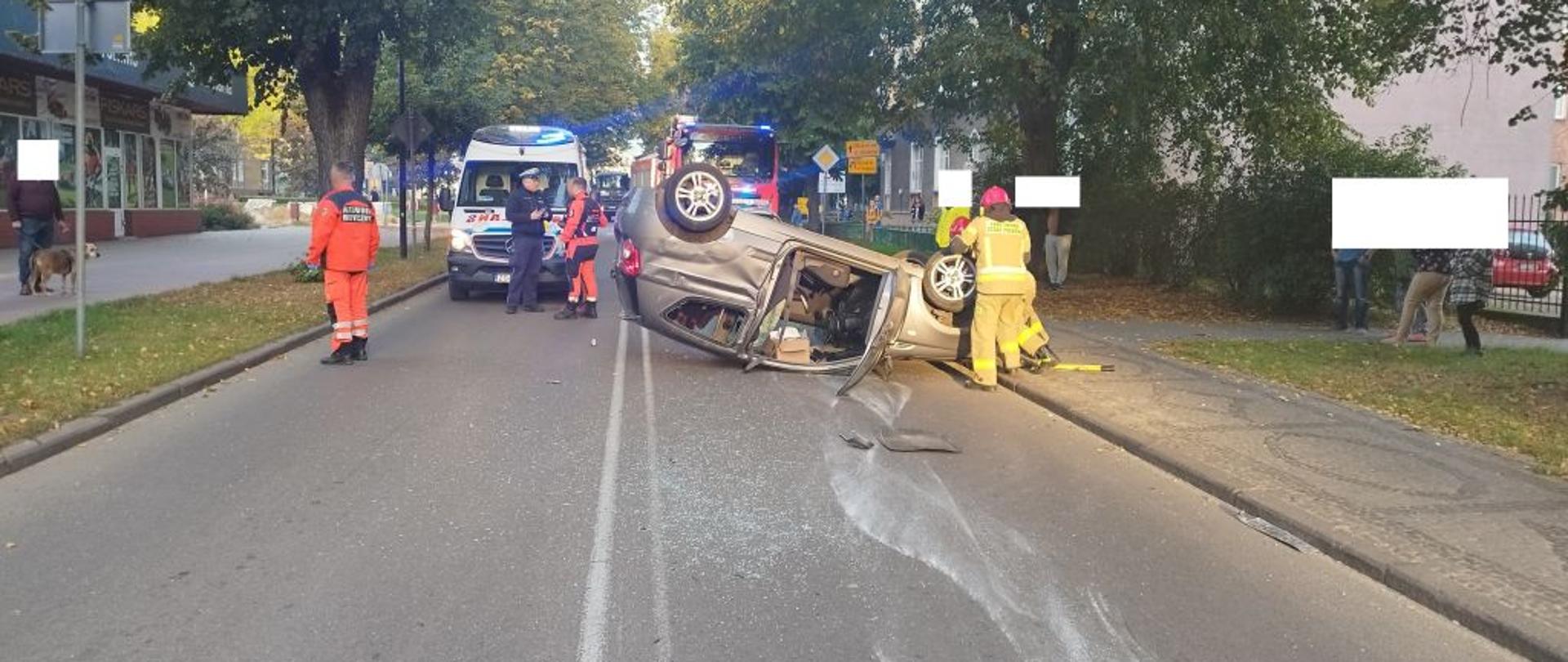 Strażacy oraz funkcjonariusze Policji znajdują się na skrzyżowaniu, gdzie doszło do kolizji. Jeden z pojazdów uderzył w słup na chodniku, co spowodowało dalsze utrudnienia w ruchu drogowym. Motocykl policyjny zabezpiecza miejsce zdarzenia, a strażacy czekają na dalsze instrukcje.