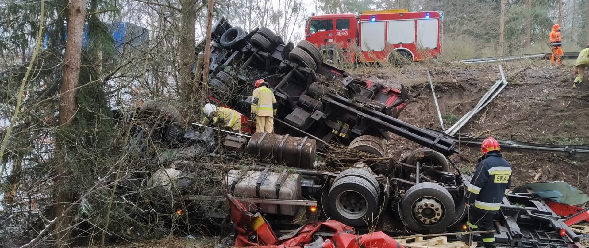 Fotografia wykonana z lasu w kierunku wraku samochodu ciężarowego leżącego na dachu, prostopadle do kierunku drogi. samochód ciężarowy leży odwrócony do góry kołami, całkowicie zniszczona jest część ładunkowa, przyczepa leży powyżej ciągnika także do góry kołami, oparta częściowo o nasyp i o drzewo, całkowicie zniszczona jest część ładunkowa, urwany zaczep. Przy wraku pracują strażacy w ubraniach specjalnych i w hełmach. Powyżej na nasypie na pasie jezdni stoi samochód pożarniczy z włączonymi niebieskimi światłami. Jest widno, dookoła leży cienka warstwa śniegu.