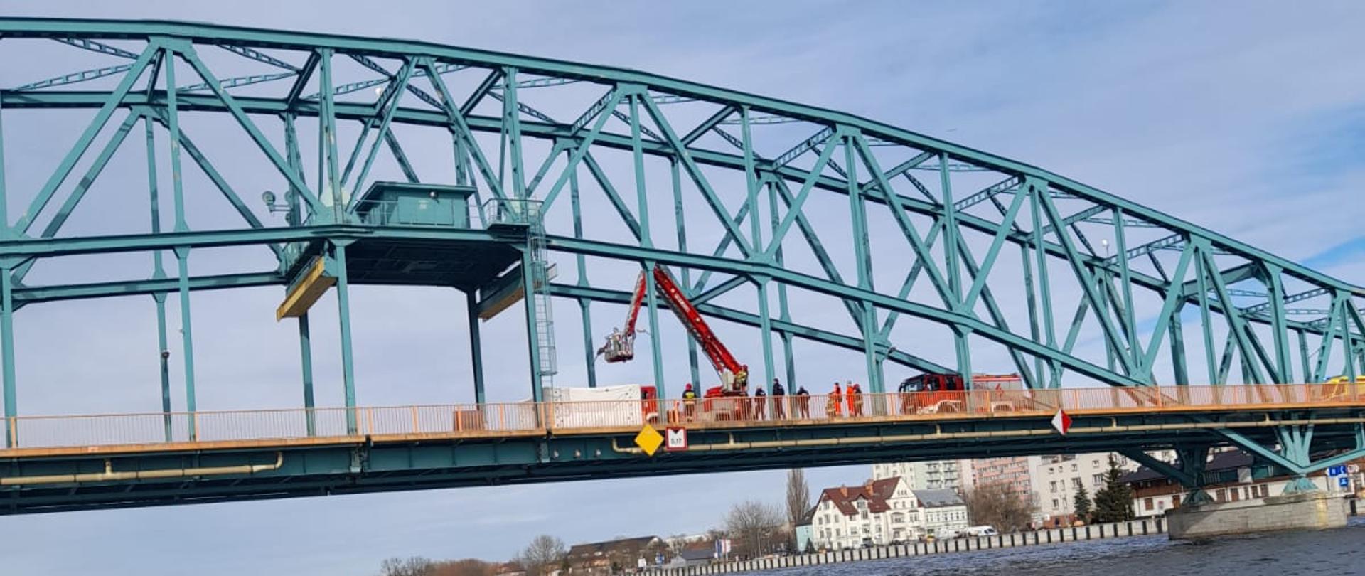 Zdjęcie przedstawia most w miejscowości Gryfino. Na moście widać rozłożony podnośnik Państwowej Straży Pożarnej. Kosz podnośnika unosi się w stronę osoby, która chce skoczyć z mostu. 