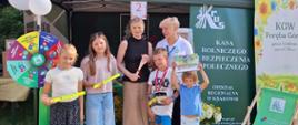 Przed stoiskiem informacyjnym KRUS podczas wydarzenia „Największa Uratowana Szarlotka w Małopolsce” stoją przedstawiciele KRUS oraz uczestnicy wydarzenia, w tym dzieci. W tle znajduje się namiot z oznaczeniami Kasy Rolniczego Ubezpieczenia Społecznego oraz materiały promocyjno-edukacyjne.