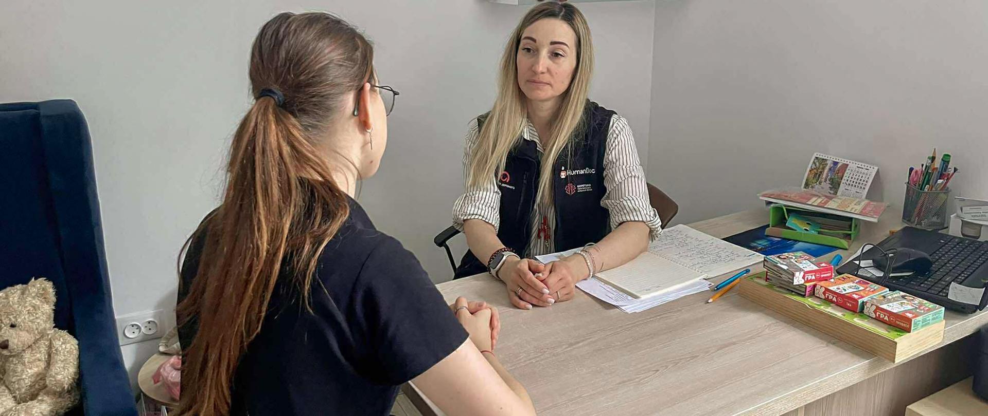 Dwie kobiety rozmawiają ze sobą przy biurku, jedna słucha drugiej i ma na sobie kamizelkę z logo Polska pomoc oraz HumanDoc