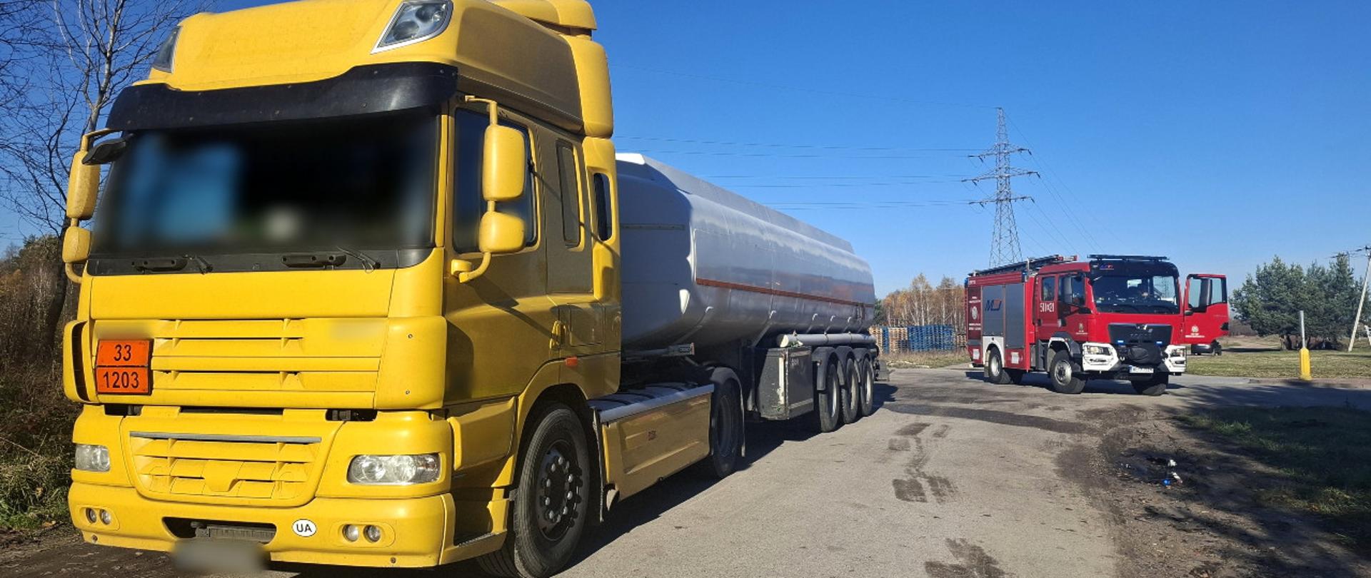 Na pierwszym planie: ciągnik siodłowy z podpiętą naczepą typu cysterna ADR. W tle: samochód gaśniczo-ratowniczy Straży Pożarnej. 