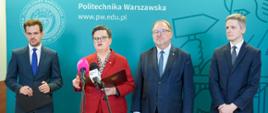 Konferencja prasowa w Politechnice Warszawskiej. Przy mikrofonach stoi Wiceminister Katarzyna Lubnauer w czerwonym garniturze, trzymając otwartą teczkę. Obok niej stoją trzej mężczyźni w ciemnych garniturach. W tle widoczna jest turkusowa ścianka z logo uczelni i adresem strony internetowej pw.edu.pl.