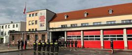 STRAŻACY ŚWIĘTUJĄ DZIEŃ FLAGI RZECZYPOSPOLITEJ POLSKIEJ. Na zdjęciu widać strażaków podczas uroczystego apelu z okazji Dnia Flagi, strażacy stoją w rzędzie, za nimi budynek strażnicy, flaga Polski uroczyście wciągana na maszt.