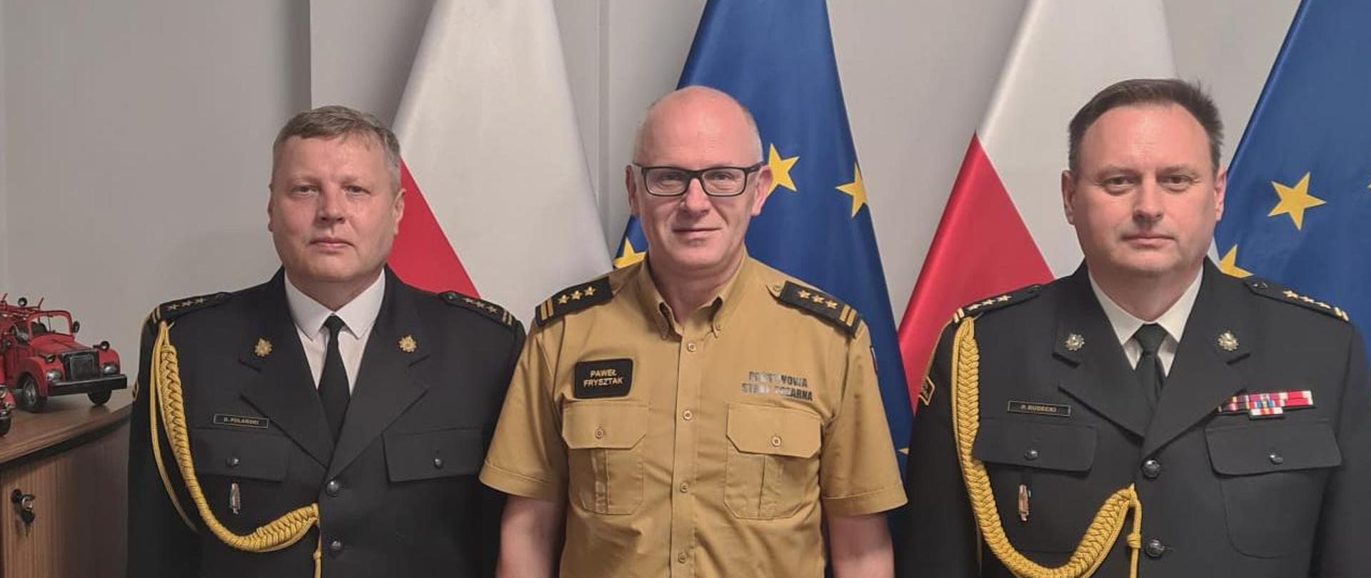 Trzech oficerów Państwowej Straży Pożarnej stoi, pozując do zdjęcia. Za nimi w tle flaga Polski i Unii Europejskiej. Na zdjęciu od lewej zastępca komendanta wojewódzkiego PSP w Łodzi, zastępca komendanta głównego PSP oraz łódzki komendant wojewódzki PSP w Łodzi.