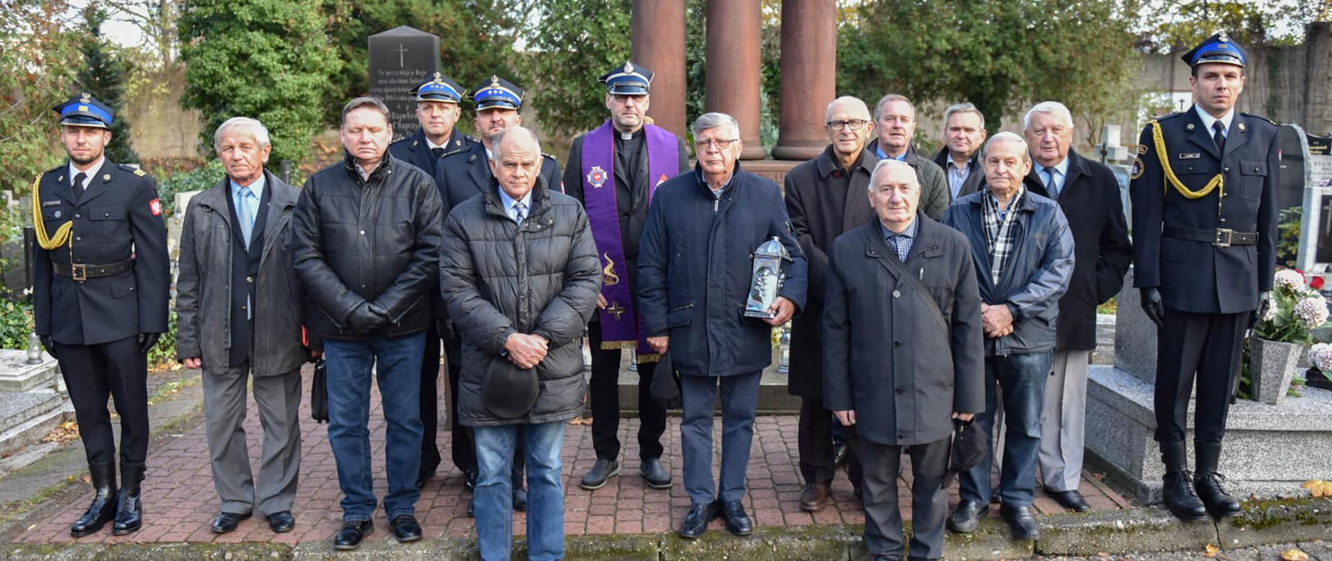 dwóch strażaków, kapelan oraz kilku mężczyzn pozują do zdjęcia na tle pomnika upamiętniającego strażaków poległych w czasie działań ratowniczo-gaśniczych