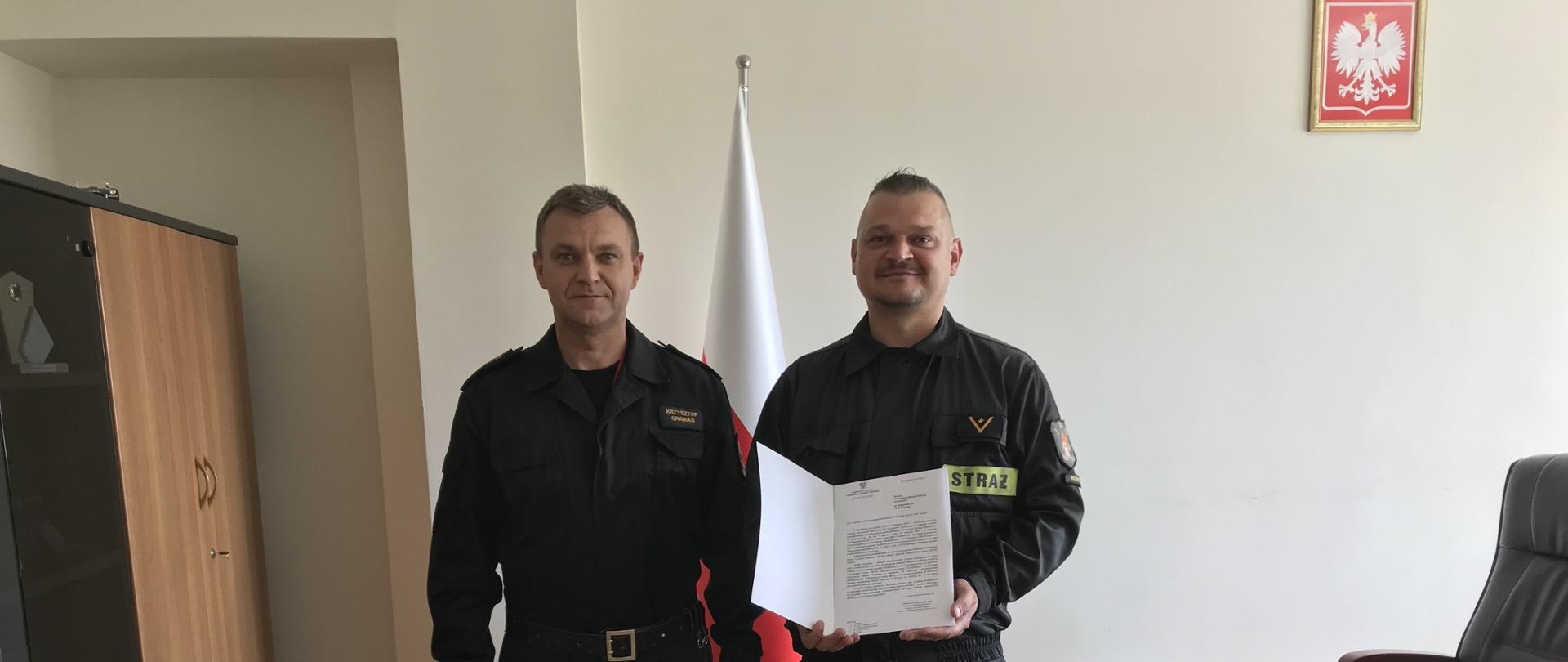 Zdjęcie zrobione w gabinecie Komendanta Powiatowego Państwowej Straży Pożarnej na tle biało-czerwonej flagi. Funkcjonariusze ubrani w czarne mundury (dowódczo-sztabowy po lewej, koszarowy po prawej). Druh prezes OSP przedstawia promesę trzymając ją w rękach.
