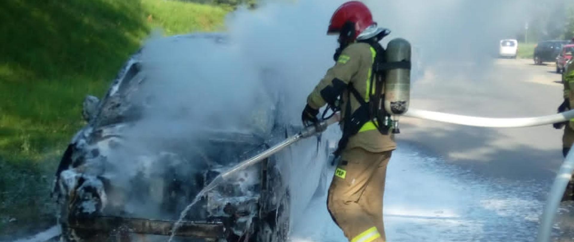 Zdjęcie przedstawia samochód osobowy, który uległ spaleniu oraz ratownika podczas akcji ratowniczo-gaśniczej.