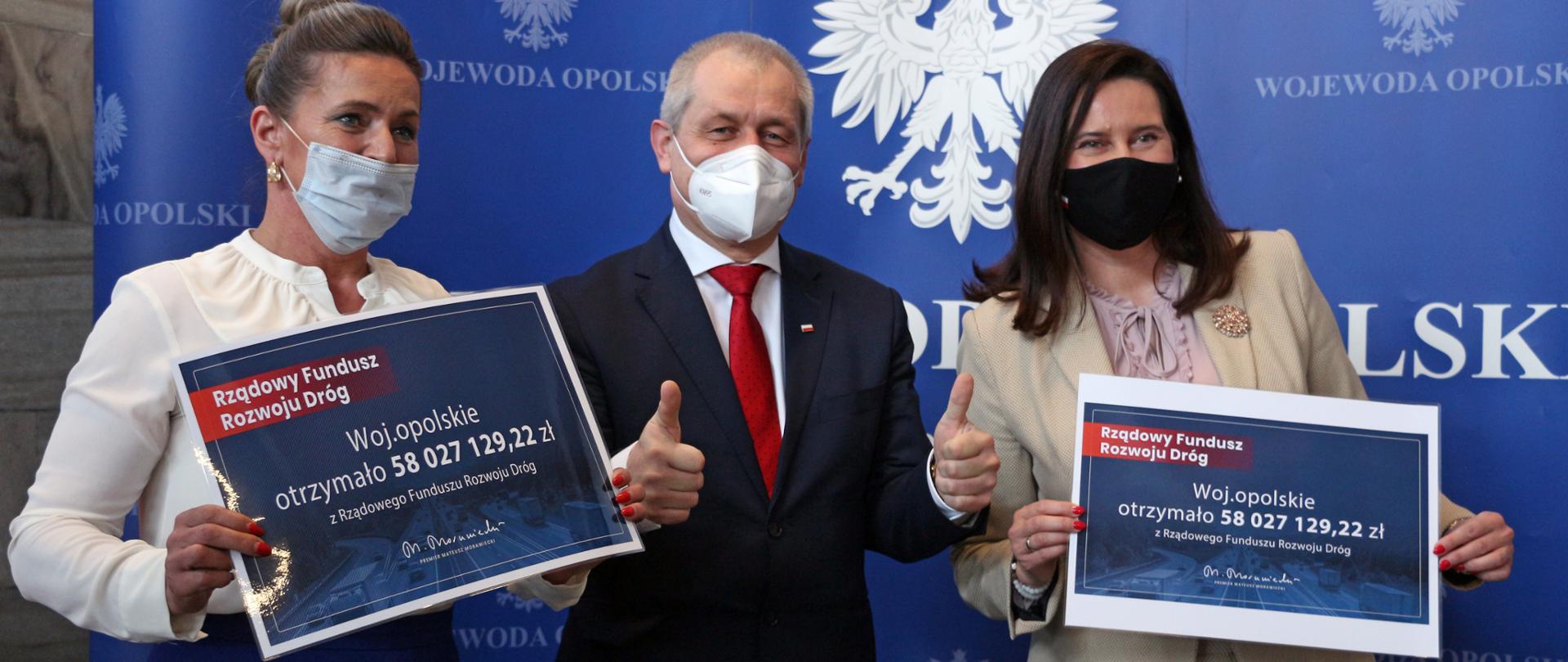 Na zdjęciu trzy osoby, stojące na tle granatowej ścianki z godłem Polski. Po lewej stronie poseł na Sejm K.Czochara, wojewoda opolski Kłosowski oraz - po prawej jego stronie - poseł na Sejm RP V.Porowska. Obie panie poseł trzymają w dłoniach czek - wydruk z informacją o przyznanej dotacji w ramach programu. Wojewoda pozuje do zdjęć z uniesionymi kciukami, na znak sukcesu. 