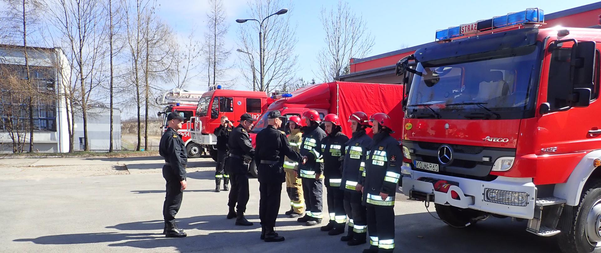 Komendant Główny oraz Komendant Wojewódzki ubrani w ubrania dowódczo sztabowe witają się ze strażakami stojącymi w szyku, Strażacy ubrani w ubrania specjalne w hełmach stoją przed samochodami pożarniczymi.