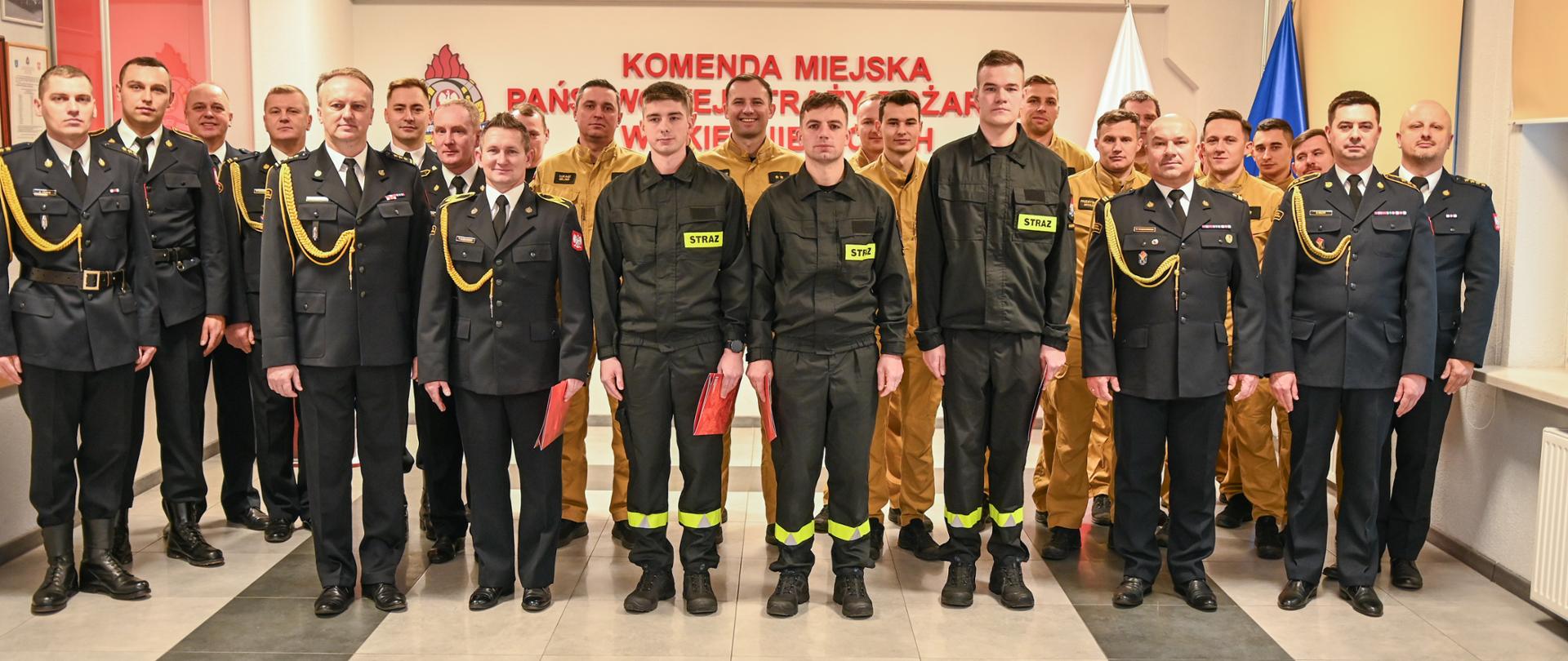 Grupowe zdjęcie funkcjonariuszy i strażaków PSP w umundurowaniu wyjściowym, ćwiczebnym i specjalnym, wykonane w sali Komendy Miejskiej PSP w Skierniewicach.