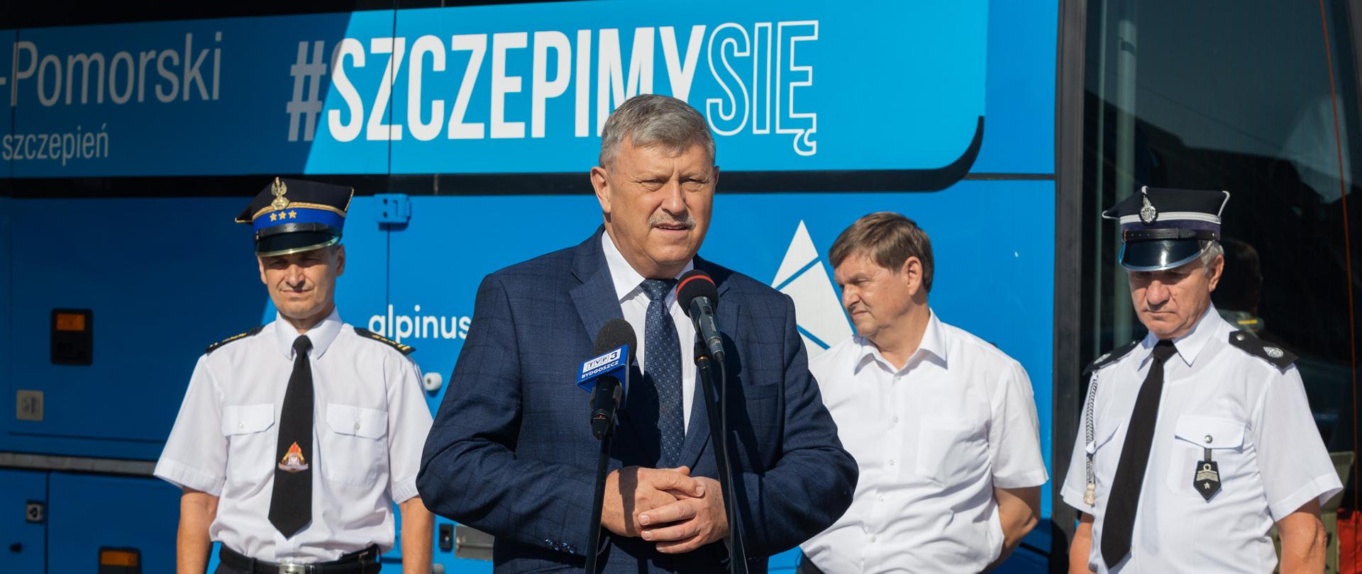 Na zdjęciu głos zabiera Wicewojewoda Józef Ramlau. W tle samorządowcy a za nim niebieski szczepionkobus.