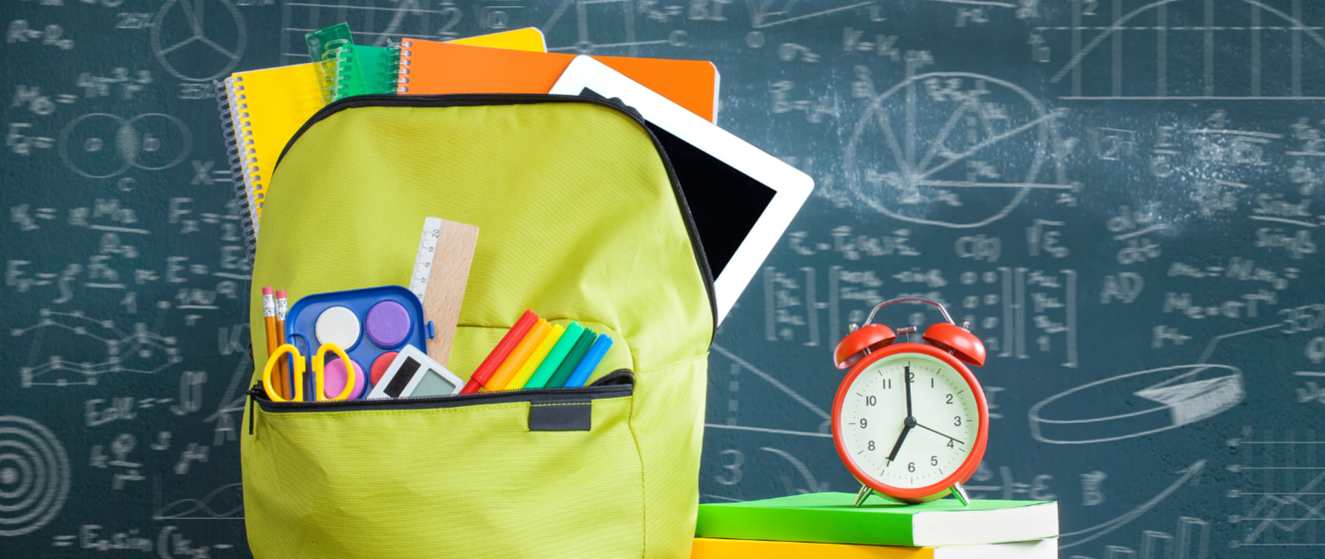 Plecak na tle zapisanej tablicy szkolnej. Plecak jasnozielony spakowany widać wystające książki i przybory szkolne.