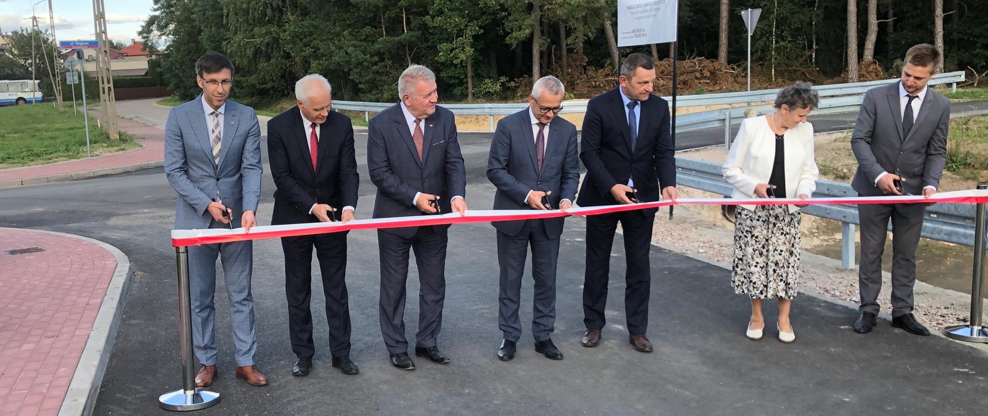Na zdjęciu znajdują się elegancko ubrani kobieta i sześciu mężczyzn. Osoby te stoją na asfalcie przebudowanej drogi, przed biało-czerwoną wstęgą. W tyle znajdują się kępy drzew. 