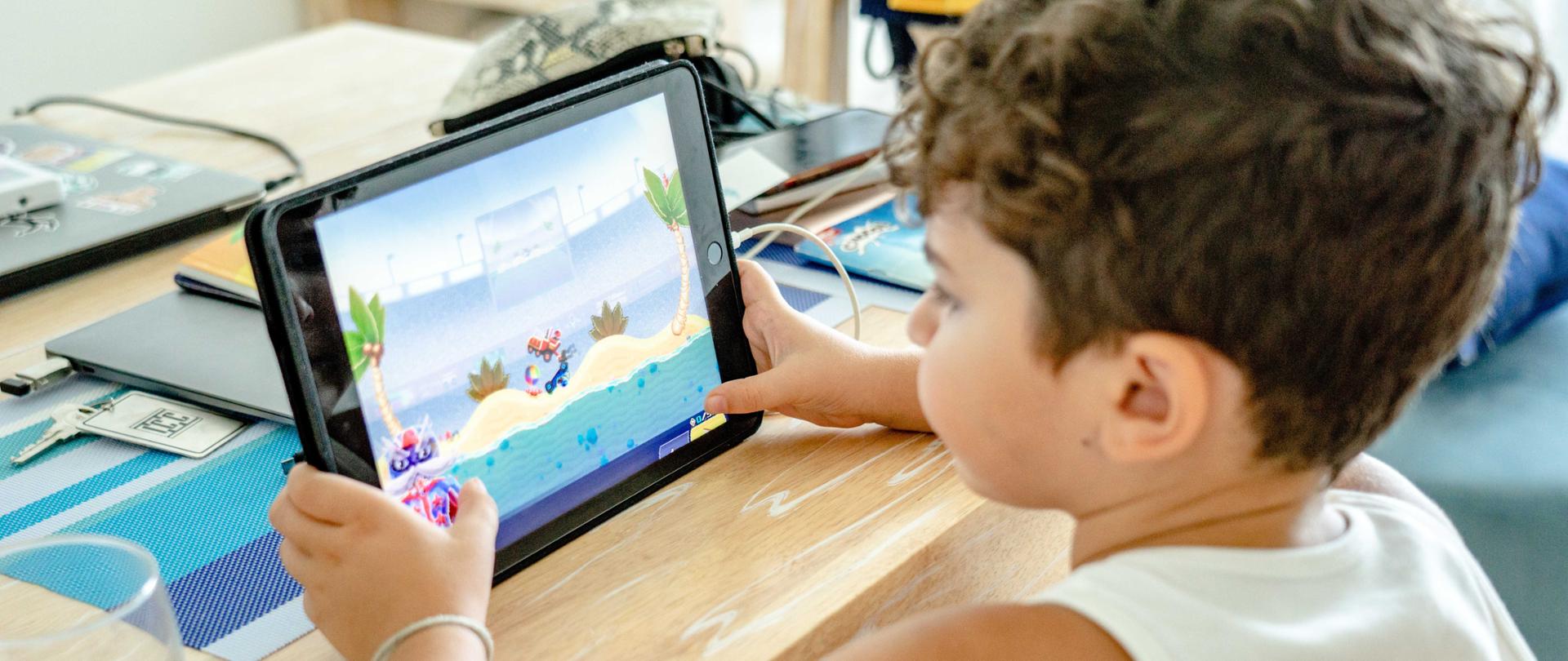 Zdjęcie w poziomie. Chłopiec ubrany w biały podkoszulek siedzi przy stole. Gra na tablecie w grę.