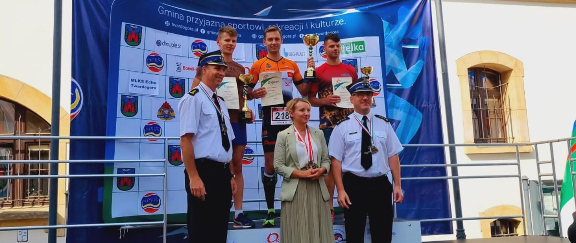 na zdjęciu widać podium, na którym stoją trzej najlepsi biegacze. Przed nimi komendanci straży pożarnej - uroczystość odznaczenia zwycięzców 