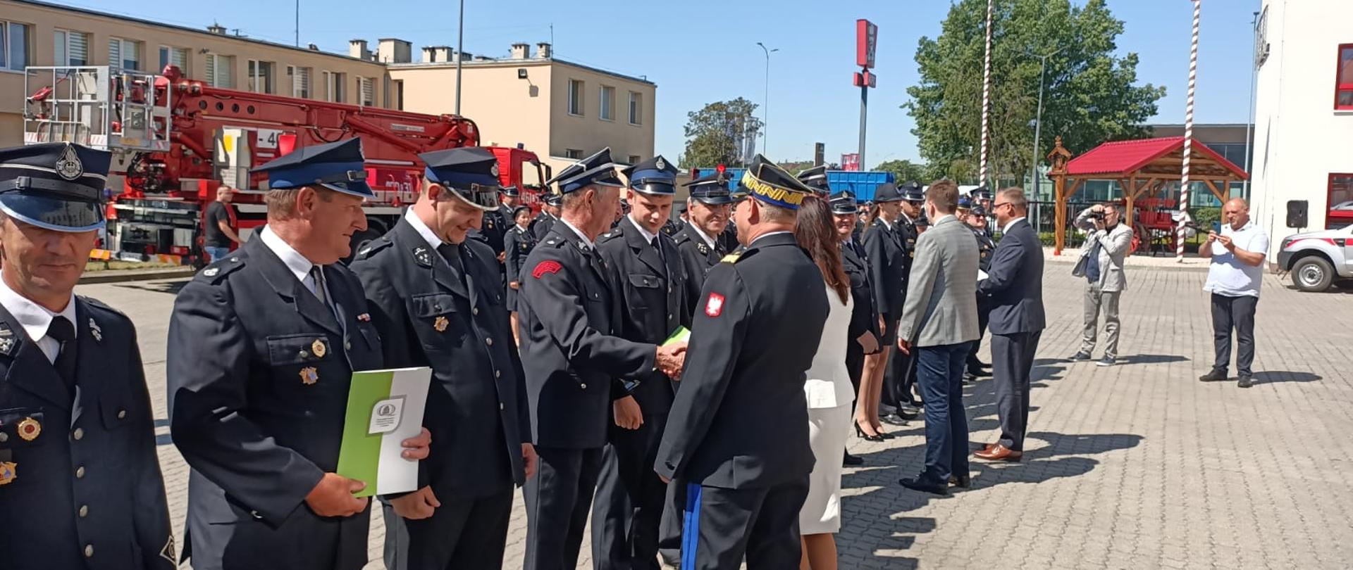 Na placu stoją w szeregu druhowie i odbierają listy gratulacyjne. Wręczają je Prezes WFOŚiGW, Minister, Pani Poseł i komendant wojewódzki. Wszyscy ubrani na wyjściowe. W tle pojazd strażacki.