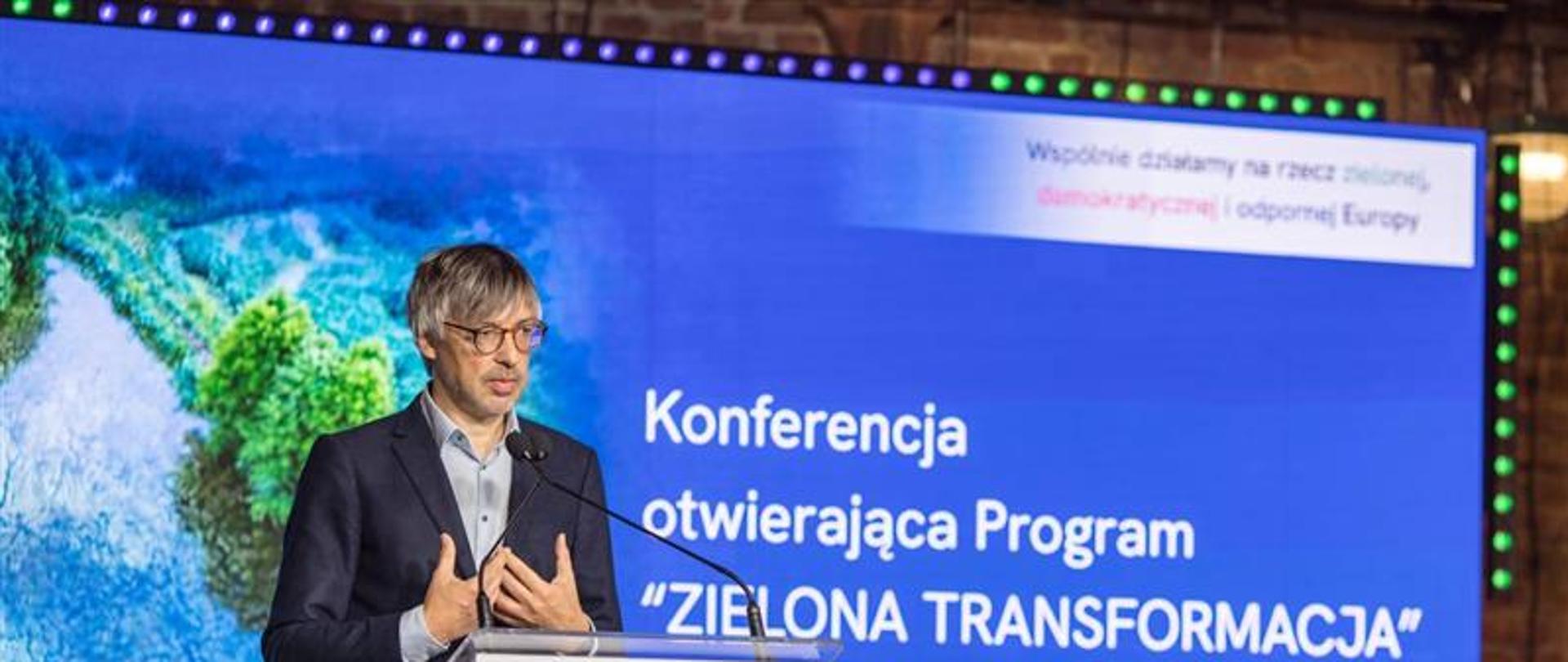 Kadr przedstawia zbliżenie na ekran i mężczyznę za mównicą, podczas konferencji otwierającej Program 'Zielona Transformacja'. Przemawia wiceminister Klimatu i Środowiska, Krzysztof Bolesta