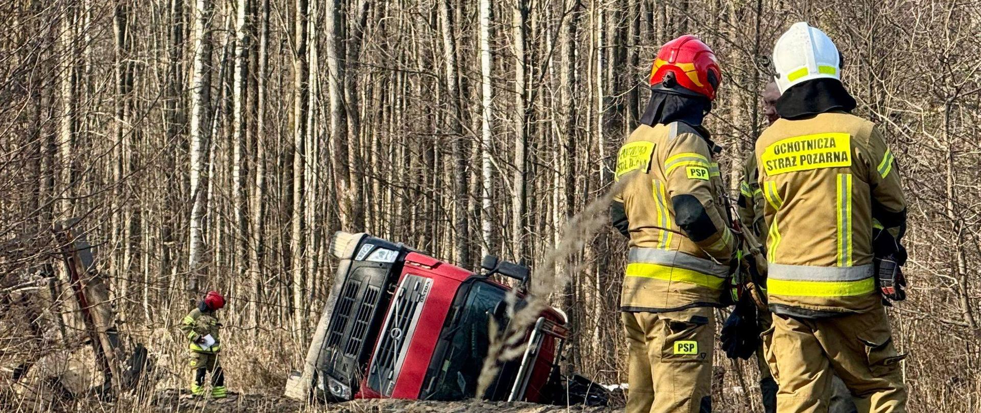Z prawej strony zdjęcia stoi dwóch strażaków w mundurach bojowych, jeden w czerwonym drugi w białym hełmie. Leśna droga, las, drzewa bez liści, słonecznie. W oddali widać czerwoną kabinę samochodu ciężarowego, leży na boku.