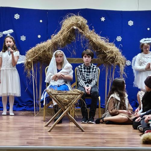 Zdjęcie przedstawia Maryję z Dzieciątkiem Jezus oraz Józefa siedzących w stajence. W tle imitacja gwiaździstego nieba. Po bokach stoją aniołki. 