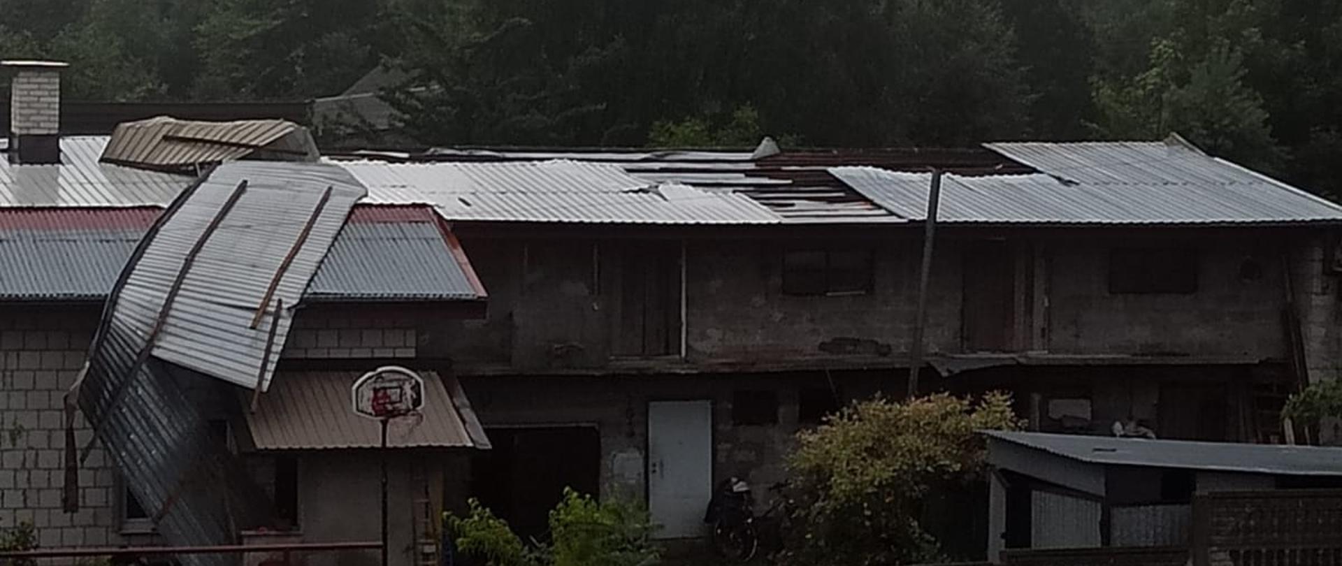 Na zdjęciu widać uszkodzony dach budynku gospodarczego. Oderwana blacha zwisa niebezpiecznie z dachu. W tle widać las.