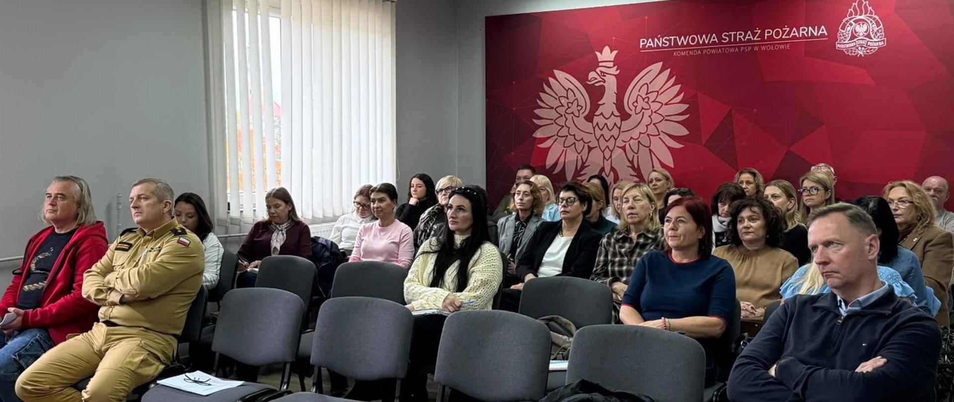 Grupa osób siedząca w sali konferencyjnej Komendy Powiatowej Państwowej Straży Pożarnej w Wołowie, słuchająca prelekcji.